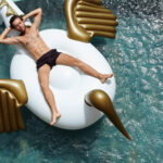 Leisure and recreation concept. Top view of young businessman with fit body lying on inflatable dragon mattress, holding hands behind his head, keeping eyes closed, enjoying long-awaited vacations